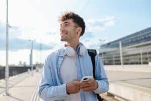 Junger Mann mit weißen Over-Ear-Kopfhörern um den Hals steht bei Tageslicht auf einem modernen Bahnsteig, schaut lächelnd zur Seite und hält ein Smartphone in der Hand. Im Hintergrund sind ein Bürogebäude und ein heller Himmel zu sehen.