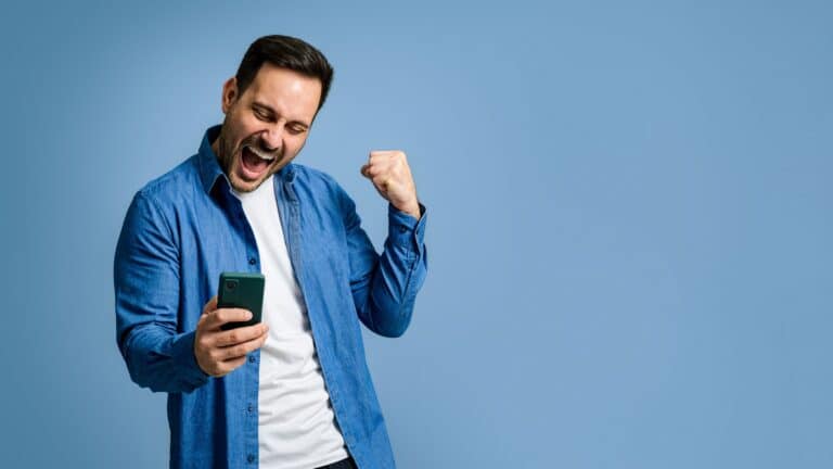 Gutaussehender mittelgroßer erwachsener Mann, gekleidet in Jeanshemd, schreiend und fröhlich die Faust ballend, während er gute Nachrichten über ein Smartphone auf blauem Hintergrund liest